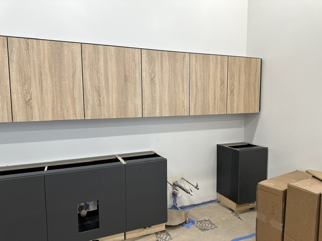 Wood-grain wall cabinets hung above dark base cabinets during a salon installation, with plumbing stubs at the wall